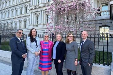 六名身着商务装的人站在华盛顿艾森豪威尔行政办公大楼外拍照, DC. 他们后面是一棵盛开的樱桃树.