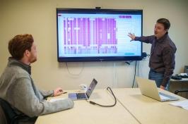 照片中，两名男子正看着屏幕上显示的表示基因组序列的粉色线条.